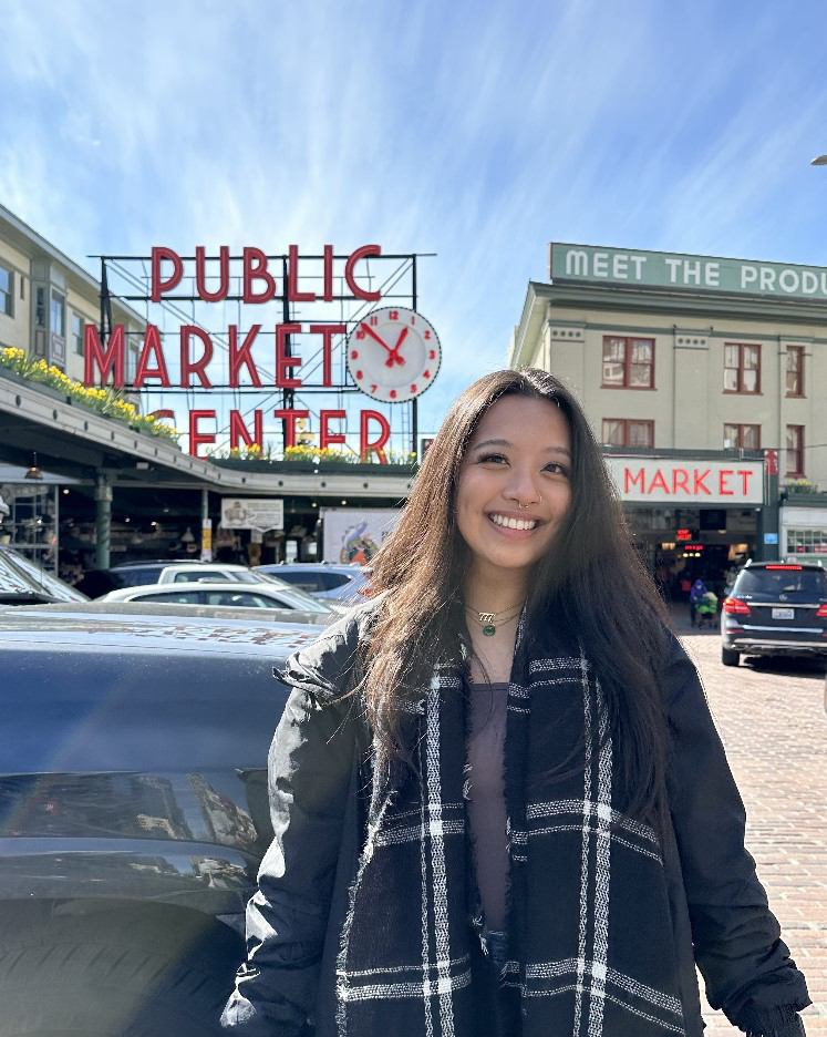This image contains a tan skin person who has long dark brown hair, dark brown eyes, wearing a black jacket, a black and white scarf, has a hoop septum piercing, and is smiling at the camera. To the left of the photo is a red sign that reads "Public Market Center" with a clock that has hour hand between the 10 and 11 and the minute hand at the 1 with parked cars.