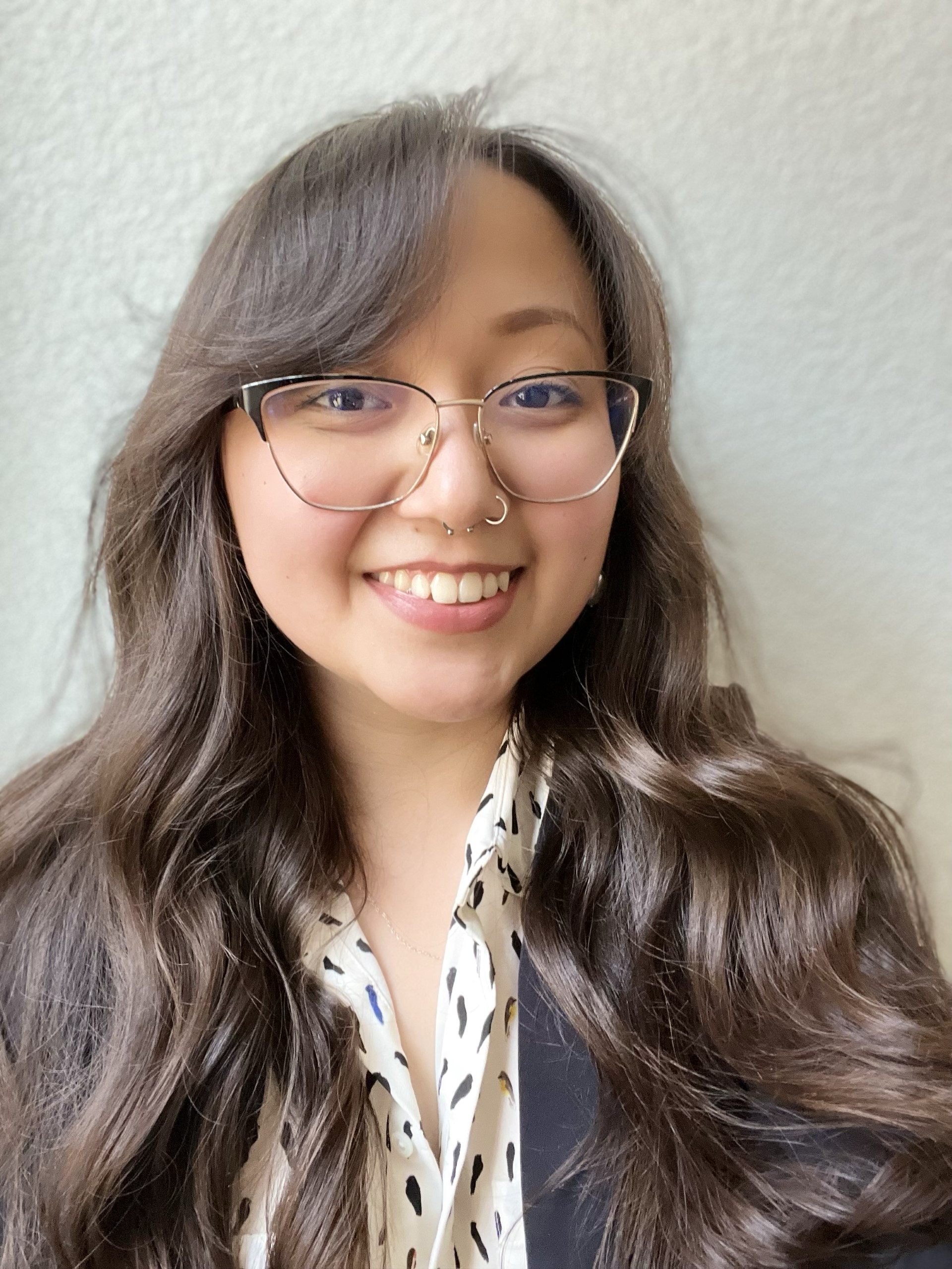This photo contains a person who is a multiracial woman with light brown skin with small shades of yellow, long brown hair, a right nostril piercing, and a septum piercing. She is wearing eyeglasses, a white bird print blouse, and a black blazer. She is smiling at the camera in front of a white background.