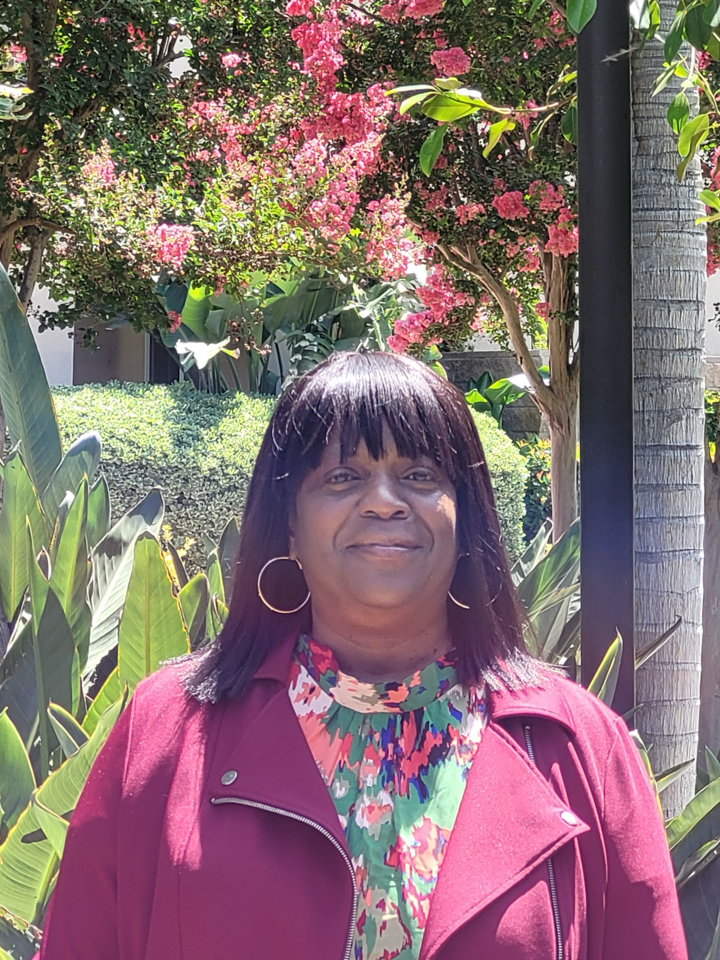 I am an African American woman with straight brown shoulder length hair with bangs. I have large brown eyes, cocoa brown skin and I am wearing hoop earrings. I'm wearing a multicolored blouse with a red jacket. Behind me are green plants including palms and red flowers.
