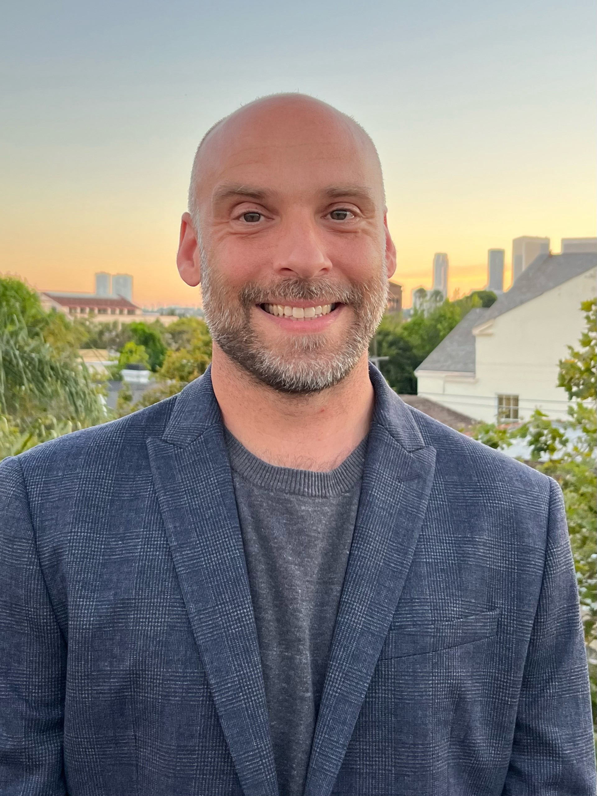 A white passing, cis, man; with a shaved head, and a trimmed, dark brown and gray beard, is smiling. He is wearing a Navy sports jacket with a gray sweater underneath it. The subject stands in front of a cityscape revealing the tops of trees with green leaves and gray shingled roofs of houses. In the far distance, skyscrapers rest against the horizon as the setting sun illuminates the sky with a pale blue, yellow, and orange.