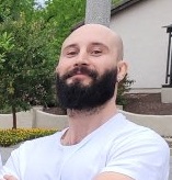 Image Description: This image contains a white, cis gender person, brown eyes, bald head, brown beard, and silver hoop earrings, wearing a white t-shirt, and in the background is a house with some green trees and yellow flowers.