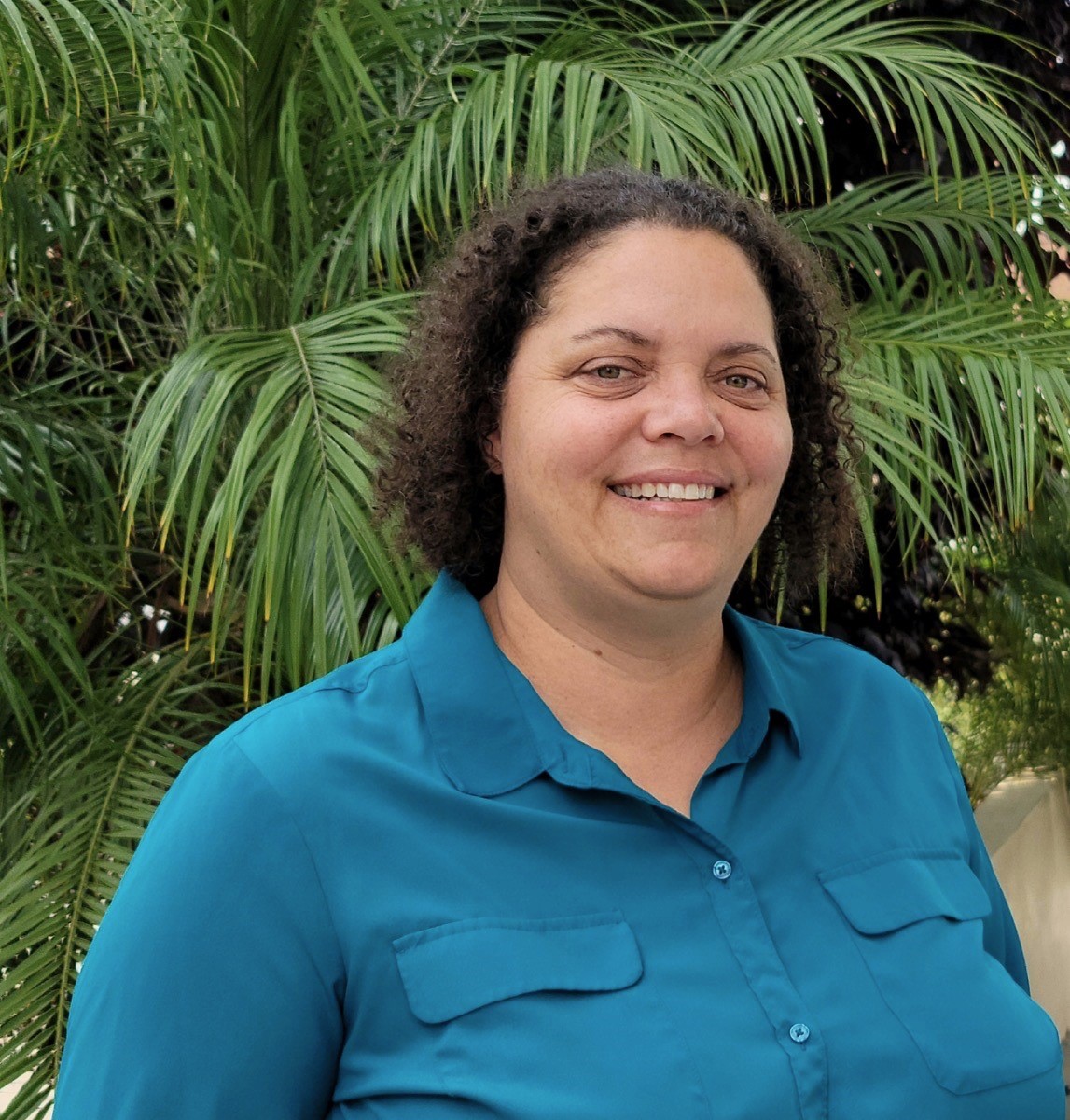 This image contains a brown curly haired person with tan olive skin and hazel eyes standing in front of a  palm tree wearing a teal button up blouse with 2 front pockets smiling while looking at the camera. 