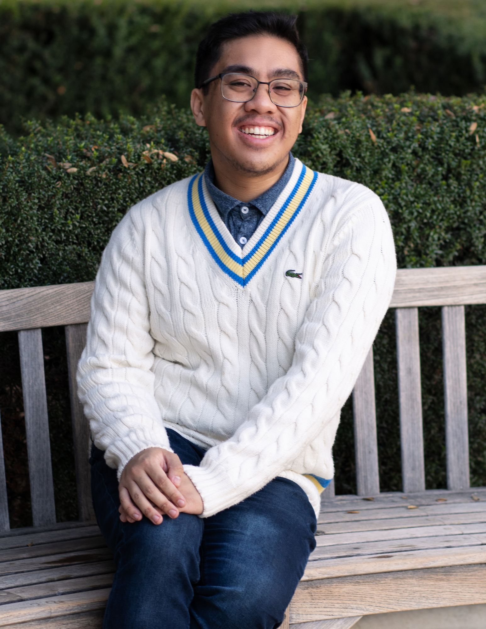 This image contains an asian-american, cis man with brown eyes, wearing glasses, with short black hair, wearing a fluffy, knitted white sweater, over a blue polo shirt, topped with a pair of dark blue jeans, sitting on a bench. In the background are rows of  hedges and bushes.  