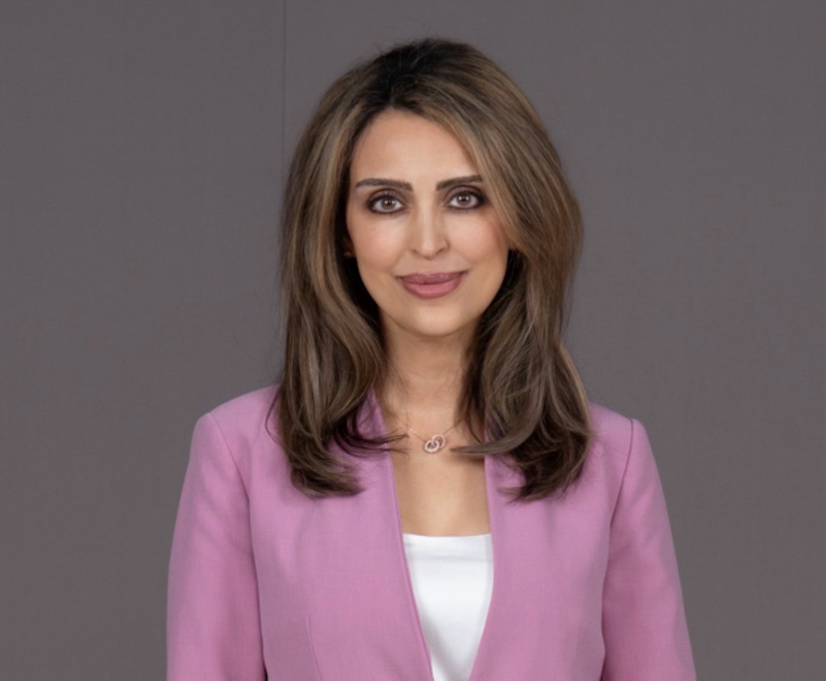 This image contains an olive-skinned person with hazel eyes and brown, straight, shoulder-length hair. The person is wearing a pink jacket, white blouse, and thin, gold necklace. The background is gray.