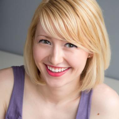 A Caucasian person with olive green eyes and shoulder-length blonde hair smiles into the camera while wearing pink lipstick and purple straps. 