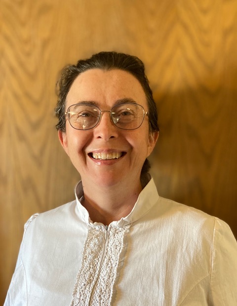 In this photo is a fifty year old person with olive skin, smiling and looking at the camera. She is wearing metal glasses, dark hair, and a white shirt with lace details. Behind her is a wooden paneled door. 