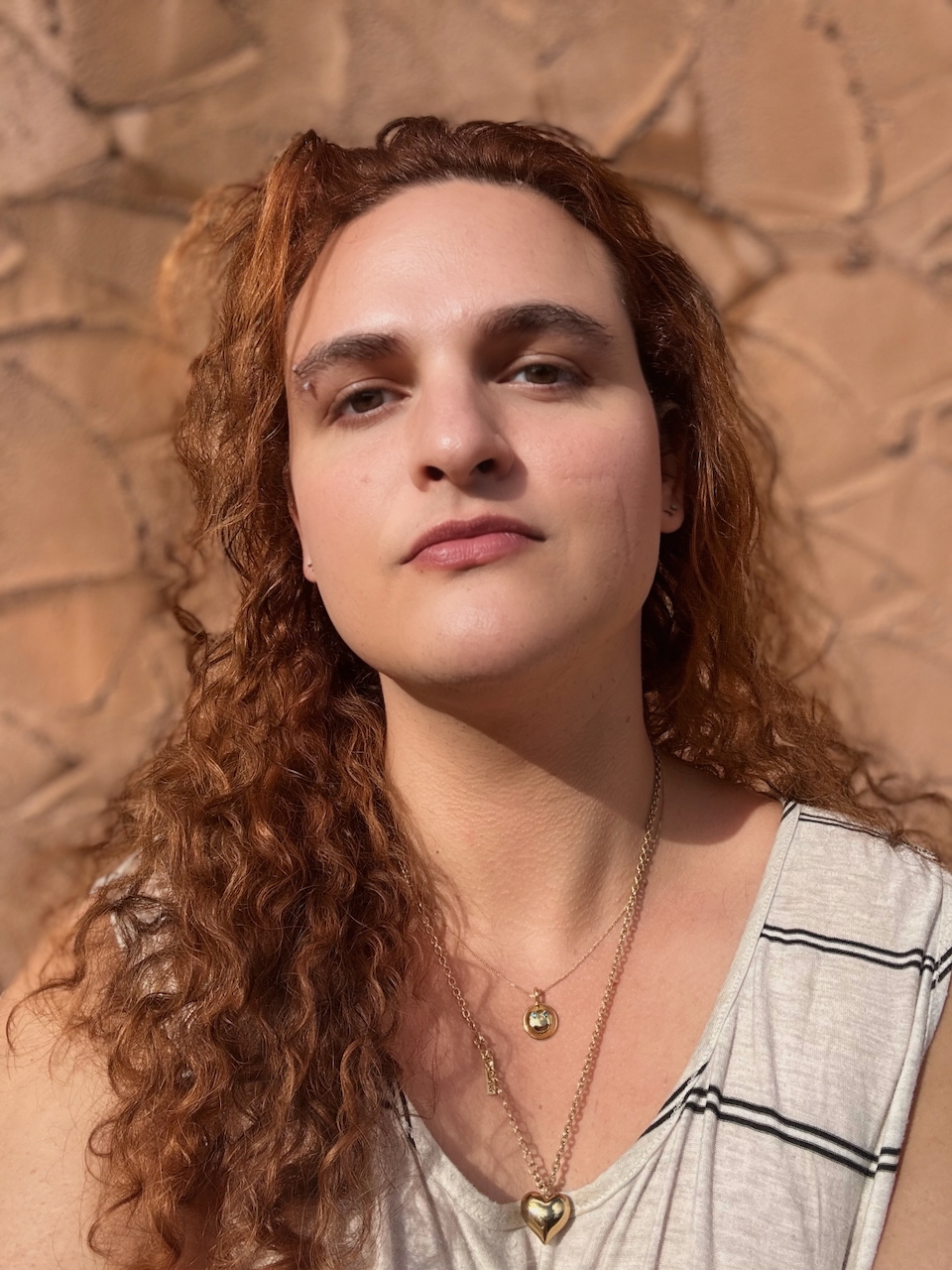 This image shows a white person with long, curly copper hair, an eyebrow piercing, and a facial scar on their left cheek. They are wearing a necklace of a gold heart and another necklace of a small, metallic smiley-face with inlaid sparkling green quartz eyes. The subject is standing against a tan cement wall, smiling and looking at the camera 